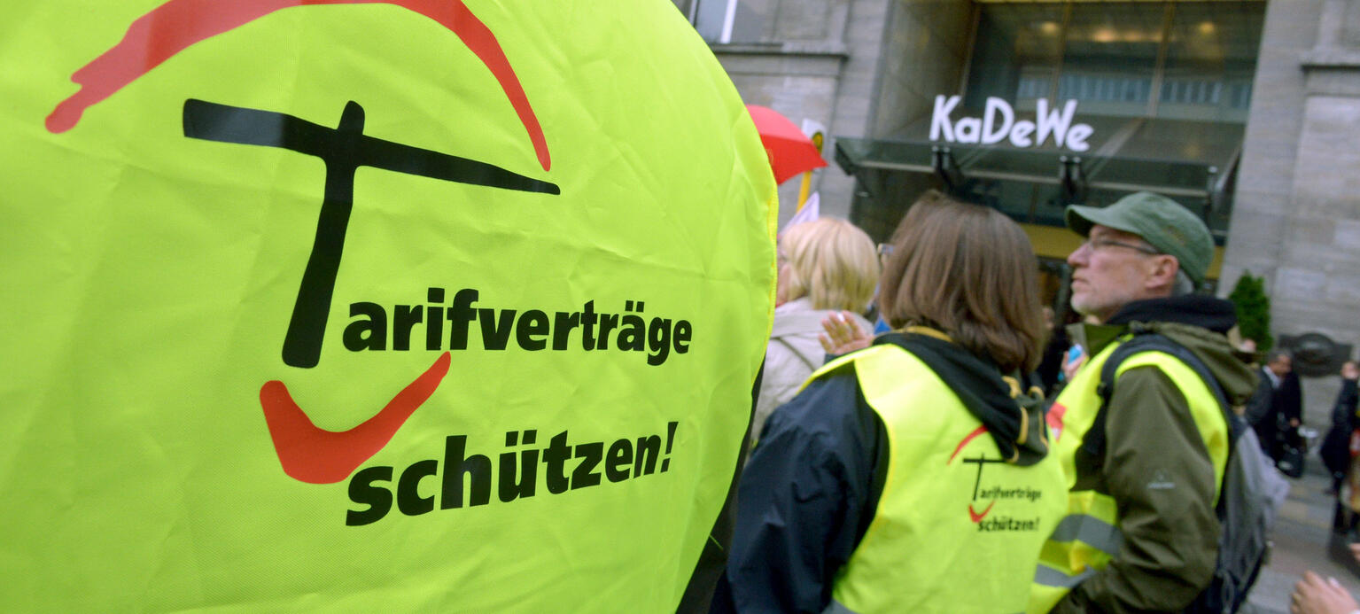 Ein Demonstrationszug läuft vor dem KaDeWe vorbei. Im Fokus steht eine gelbe Warnweste auf der 'Tarifverträge schützen!' in schwarzer Druckschrift steht.