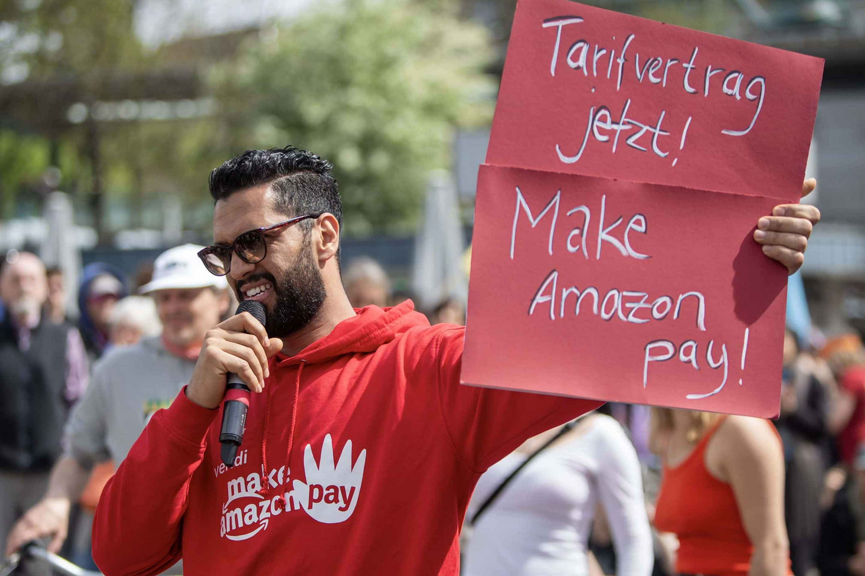 Hedi Tounsi, Betriebsrat bei Amazon, hält ein Schild hoch mit der Aufschrift: Tarifvertrag jetzt! Make Amazon pay!